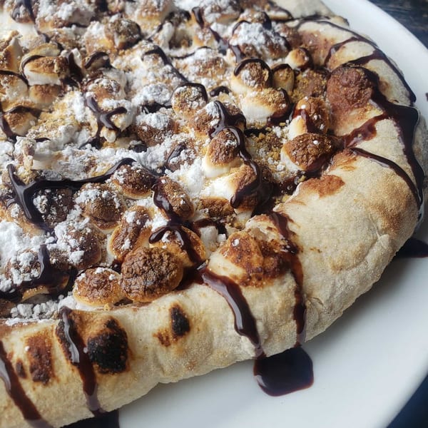 Dessert pizza with toasted marshmallows, powdered sugar, and chocolate drizzle on a white plate.