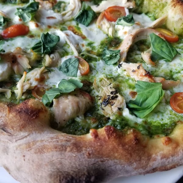 Close-up of a wood-fired pesto chicken pizza with mozzarella, cherry tomatoes, and fresh basil.